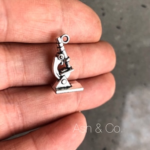 Op de afbeelding: Zilveren bedel in de vorm van een microscoop. De bedel heeft een lus aan de bovenkant om aan een ketting of armband te bevestigen.