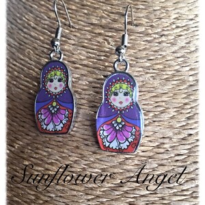 May include: A pair of silver dangle earrings with a purple, red, and white Russian nesting doll design. The dolls have a yellow face with black eyes and a pink mouth.