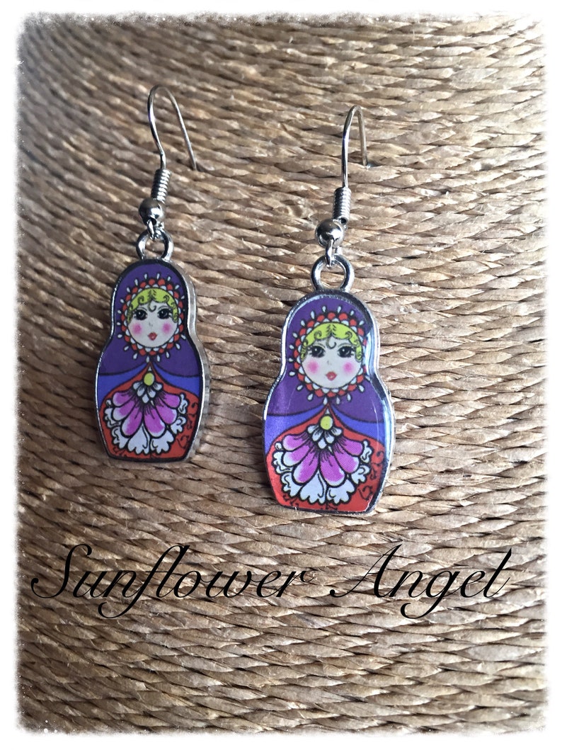 May include: A pair of silver dangle earrings with a purple, red, and white Russian nesting doll design. The dolls have a yellow face with black eyes and a pink mouth.
