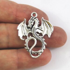 May include: Silver-toned metal dragon pendant with outstretched wings and a curled tail. The dragon is detailed with scales and has a loop at the top for attaching to a chain or cord.