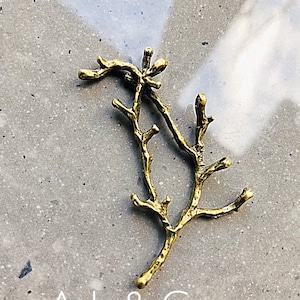 Op de afbeelding: Gouden metalen ornament in de vorm van een tak. Het ornament is een gestileerde weergave van een tak met meerdere vertakkingen. Ash & Co.