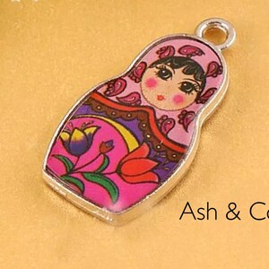 May include: A silver charm featuring a pink and purple Russian nesting doll with a floral design. The doll has a painted face with black hair and brown eyes.