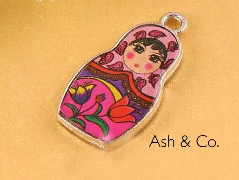 May include: A silver charm featuring a pink and purple Russian nesting doll with a floral design. The doll has a painted face with black hair and brown eyes.