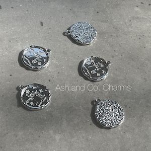 May include: Five silver-colored charms. Three are round with a decorative edge and a detailed design in the center. Two charms have a textured surface. The text "Ash and Co. Charms" is visible.