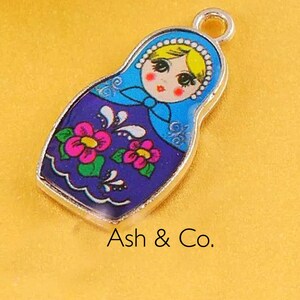 May include: A silver charm in the shape of a Russian nesting doll. The doll is painted with blue, purple, pink, yellow, and green colors. The doll has a smiling face and a floral pattern.