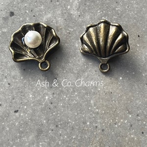 May include: Two antique gold-toned metal seashell charms with a loop for hanging. One charm has a white pearl in the center.