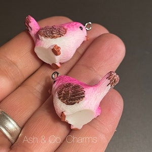May include: Two pink bird charms with brown accents and silver jump rings. The birds are facing opposite directions.
