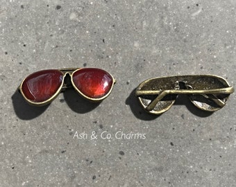 May include: A pair of gold-tone aviator sunglasses with red lenses. The sunglasses are shown from the front and back. Ash & Co. Charms is printed below the sunglasses.