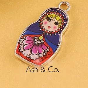 May include: A silver charm in the shape of a Russian nesting doll. The doll is painted with a blue, red, and white floral pattern. The doll's face is painted with a pink blush and a red mouth.