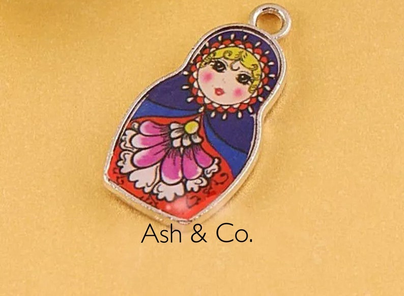 May include: A silver charm in the shape of a Russian nesting doll. The doll is painted with a blue, red, and white floral pattern. The doll's face is painted with a pink blush and a red mouth.