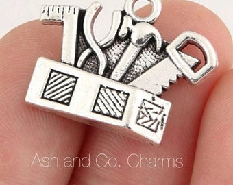 May include: Silver-toned charm featuring miniature carpenter tools, including a saw, hammer, and ruler, arranged on a rectangular base. The charm has a loop for attaching to jewelry or crafts. The text "Ash and Co. Charms" is visible.