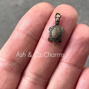 May include: A miniature, antique bronze turtle charm with a detailed shell pattern. It features a loop for attaching to jewellery or crafts. The text "Ash & Co. Charms" is visible.