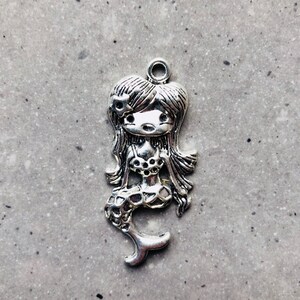 Könnte beinhalten: Silberner Anhänger einer Meerjungfrau mit einer Blume im Haar. Die Meerjungfrau trägt einen Muschelbh und hat einen Fischschwanz.