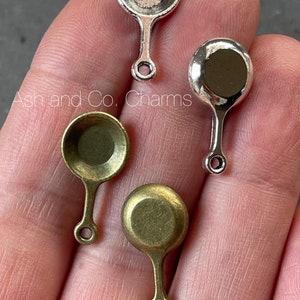Puede incluir: Cuatro encantos de sartén miniatura, dos plateados y dos de latón, con un bucle al final del mango. Ash and Co. Charms.