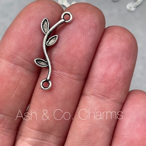 Op de afbeelding: Zilverkleurige metalen bedel in de vorm van een wijnstok met drie bladeren. De bedel heeft twee springringen om aan sieraden te bevestigen.