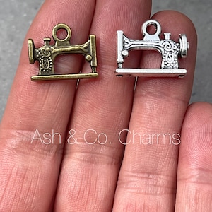 Könnte beinhalten: Zwei antike silberne Nähmaschinen-Anhänger. Ein Anhänger ist silberfarben und der andere bronzefarben.
