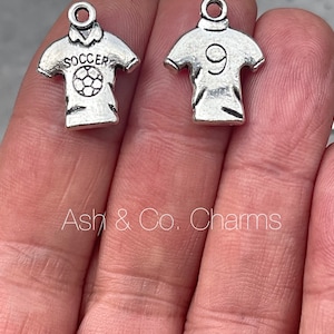 May include: Two silver charm pendants shaped like football jerseys. One jersey has the word "SOCCER" printed on it and a football graphic. The other jersey has the number "9" printed on it.