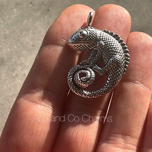 Pode incluir: Encanto de pingente de camaleão de prata com um design texturizado detalhado. O encanto tem a forma de um camaleão enroscado com a cauda enrolada ao redor do seu corpo.