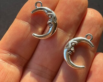 May include: Two silver-colored crescent moon charms with a face carved into each. The charms have a loop at the top for attaching to jewelry or other crafts. The moons are in a hand.