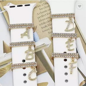 Könnte beinhalten: Weißes Apple Watch Armband mit goldfarbenen Metall-Anhängern mit den Buchstaben A, B, C, D, E und F. Jeder Anhänger ist mit klaren Strasssteinen verziert.