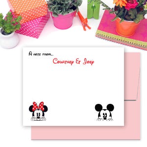 May include: A white card with a black border and a pink envelope. The card has the text "A note from... Courtney & Joey" in black text. The card also has a black and white image of Mickey Mouse and Minnie Mouse peeking over the top of the card.