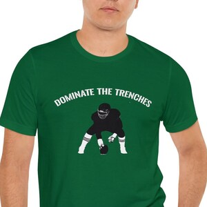 Könnte beinhalten: Ein grünes T-Shirt mit einer Silhouette eines Footballspielers in einer Drei-Punkte-Haltung und dem Text "DOMINATE THE TRENCHES".