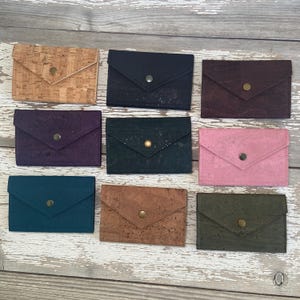 May include: A collection of nine rectangular envelope-style wallets made from cork material in various colors including natural tan, black, dark purple, deep teal, dark brown, pink, and olive green, each featuring a single metallic snap button closure.