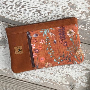 May include: A rectangular, zippered pouch with a floral pattern and a brown leather-like exterior. The floral pattern features orange, pink, and blue flowers on a rust-colored background. A small square patch with a bird logo is on the left side.