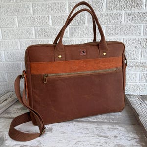 May include: A brown canvas briefcase featuring a horizontal textured leather-like strip across the front, a zippered pocket, two top handles, and an adjustable shoulder strap. A small square logo with a bird icon is centered near the top.