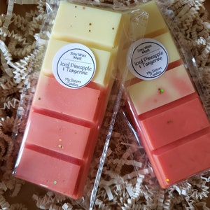 May include: Two soy wax melt bars in clear packaging, labeled "Iced Pineapple & Tangerine" by My Sisters Candles. Each bar is a gradient of yellow to coral, with gold and green glitter accents. The bars rest on white paper filler.