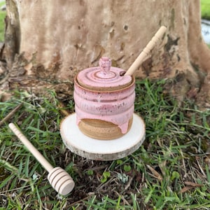 May include: A handmade ceramic honey pot with a lid and a wooden honey dipper. The pot is a light pink color with a brown base and a matching lid. The honey dipper is light brown and rests on the side. The pot sits on a small wooden slice.