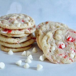 May include: A stack of soft, round cookies with white chocolate chips and red candy pieces. Several cookies are arranged nearby, with scattered white chocolate chips. The cookies have a light brown color.