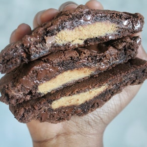 May include: Three chocolate cookies, cut in half, are stacked to reveal a peanut butter filling. The cookies are dark brown with visible chocolate chips. The peanut butter filling is a light tan color, creating a contrast.