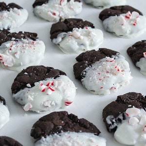 May include: Close-up of chocolate cookies dipped in white chocolate and sprinkled with crushed peppermint candies. The cookies are arranged on a white surface, with some cookies partially visible. The cookies are a festive treat.