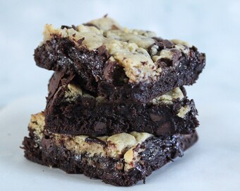 May include: A stack of three chocolate brownies with a cookie dough topping. The brownies are dark brown with visible chocolate chips and a soft, moist texture. The cookie dough topping is a light tan color.