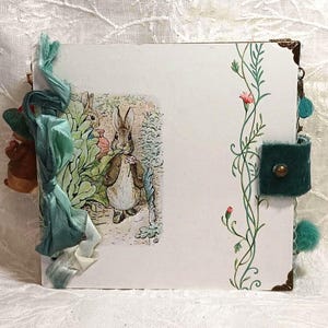 May include: A handmade scrapbook with a Peter Rabbit illustration on the cover. The cover is decorated with green and blue ribbons, a green velvet patch, and a floral design.