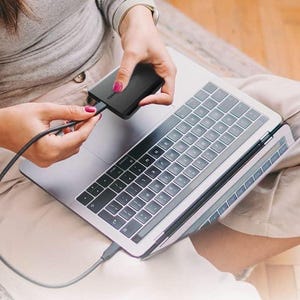 Könnte beinhalten: Eine schwarze externe Festplatte ist mit einem silbernen Laptop mit einem schwarzen Kabel verbunden. Der Laptop ist geöffnet und die Tastatur ist sichtbar. Die Person hält die Festplatte und das Kabel.