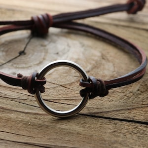 May include: A brown leather bracelet with a silver ring in the center. The bracelet is tied with a knot on each side of the ring.