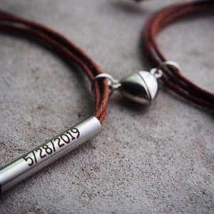 May include: Two brown leather bracelets with a silver heart charm and a silver bar engraved with the date "5/28/2019".