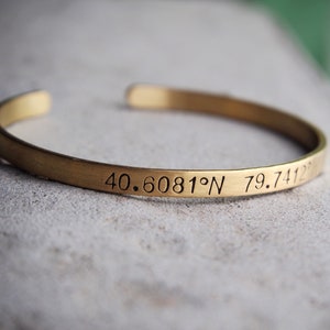 May include: A gold-colored cuff bracelet with engraved coordinates. The bracelet has a simple, open design and a brushed metal finish. The coordinates are engraved in a classic font.