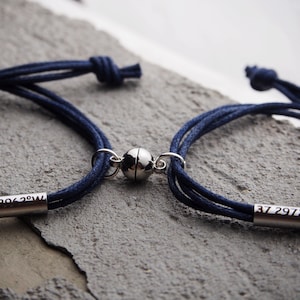Op de afbeelding: Twee donkerblauwe koordarmbanden met zilveren magneetsluitingen. De ene armband heeft de coördinaten "37 2972°N" gegraveerd op een zilveren balk, de andere heeft "78 2063°W" gegraveerd.