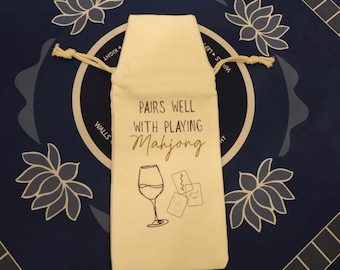 May include: A cream-colored fabric wine bottle gift bag with a drawstring closure. The front features black and gold text that reads PAIRS WELL WITH PLAYING Mahjong, accompanied by a line art illustration of a wine glass and three mahjong tiles.