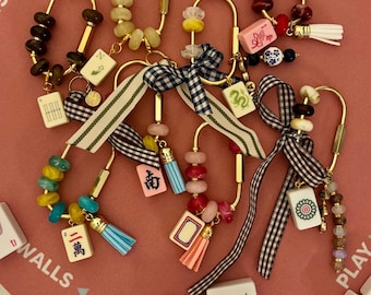 May include: A collection of gold-toned metal carabiner bag charms, each featuring a unique arrangement of colorful beads, fabric ribbons in gingham or striped patterns, small tassels, and individual mahjong tiles with various symbols and characters.