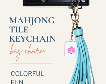 May include: A keychain featuring a white mahjong tile with a pink and blue orchid graphic, attached to a light blue leather tassel. The text reads: MAHJONG TILE KEYCHAIN bag charm COLORFUL FUN LIGHTWEIGHT. It hangs from a black hook.