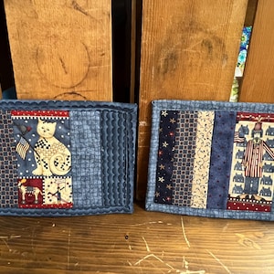 May include: Two patriotic quilted placemats. One features a cat holding an American flag, the other a figure in a striped coat. Both have blue borders and red, white, and blue fabric strips.