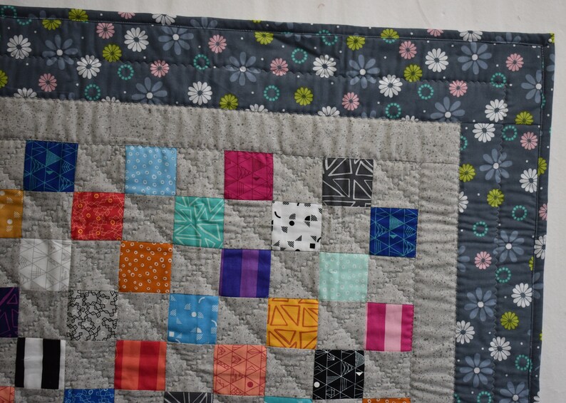 May include: A patchwork quilt with a gray background and colorful squares. The quilt is edged with a gray fabric with a white daisy print.