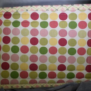 May include: A white fabric cushion with a polka dot pattern in shades of pink, yellow, and green. The cushion has a decorative border with a floral design.