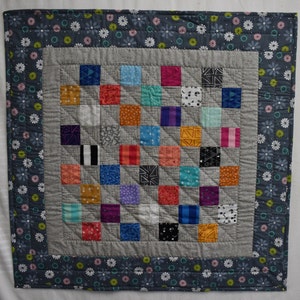 May include: A gray and white quilt with a patchwork design. The quilt is made up of small squares of different colors and patterns. The quilt is surrounded by a border of gray fabric with white flowers.