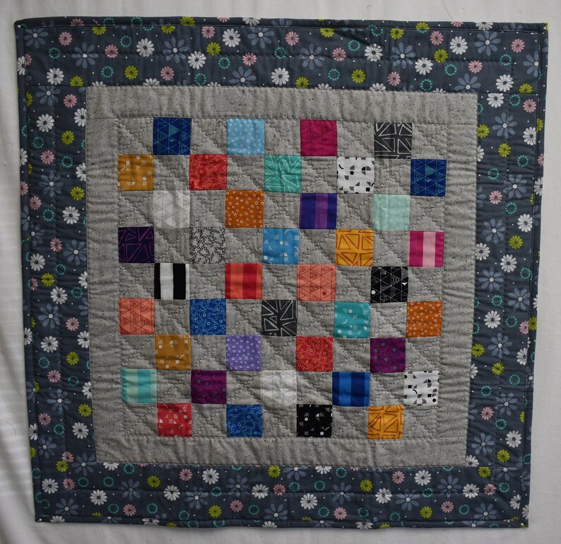 May include: A gray and white quilt with a patchwork design. The quilt is made up of small squares of different colors and patterns. The quilt is surrounded by a border of gray fabric with white flowers.