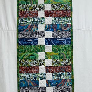 May include: A colorful patchwork quilt with a variety of patterns and colors, including green, blue, red, and white. The quilt is made up of horizontal strips of fabric, with a white square in the center of each strip. The quilt is framed with a green border.